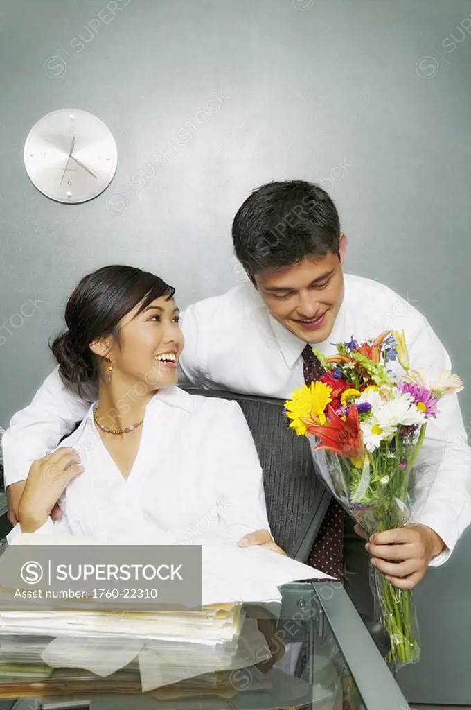 Businesswoman at desk covered in paperwork, attractive man in suit bringing her flowers