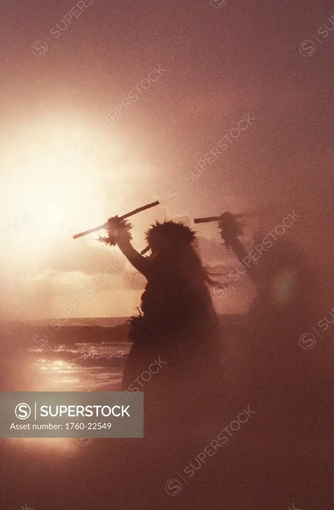 Pair of hula dancers at sunset, holding sticks, soft focus blurry background