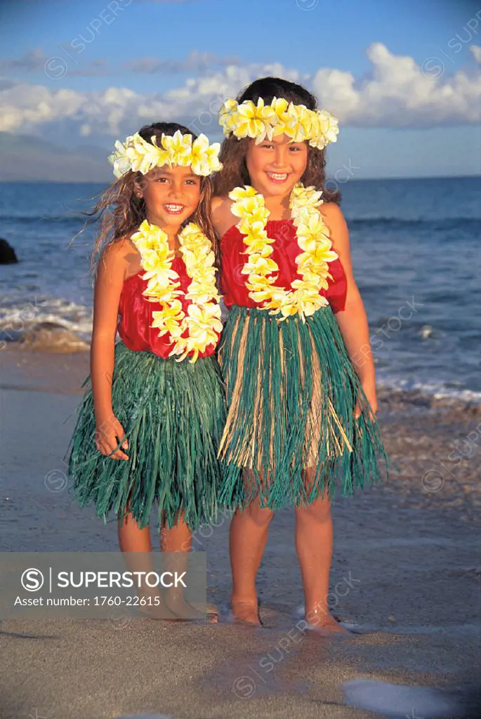 Two smiling girls on beach in hula costume, ti leaf skirt, red top, plumeria lei