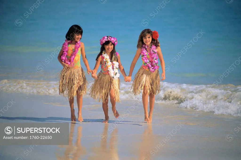 Three young girls walk on beach in water, hold hands, grass skirts, haku & lei