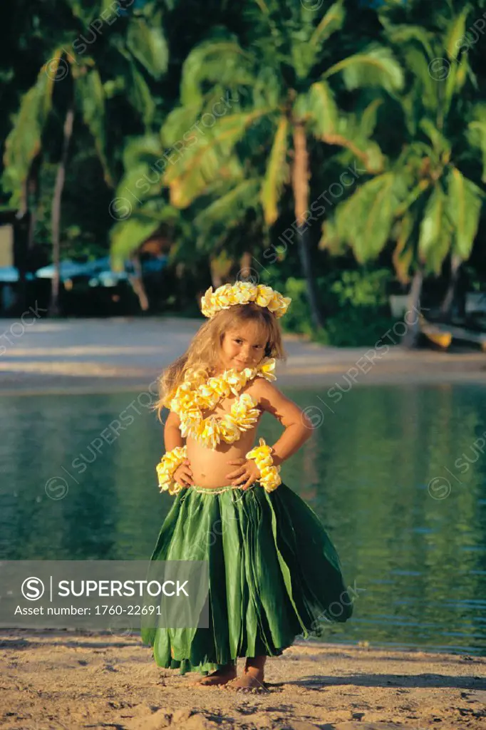 Keiki hula little girl in ti leaf skirt & plumeria leis, hands on hips, beach