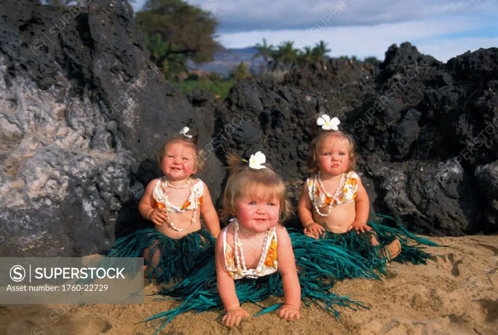 Three babies wearing hula sitting on sand with tropical rock background