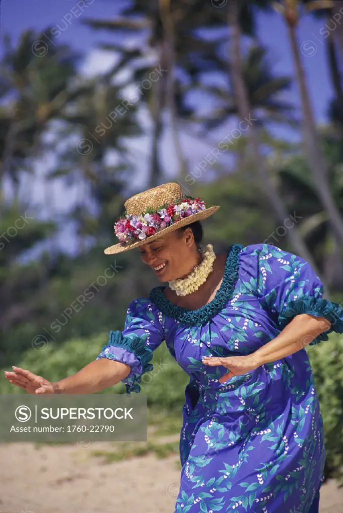 Side angle of smiling woman dances hula in muumuu wearing hat beach w/ palms background
