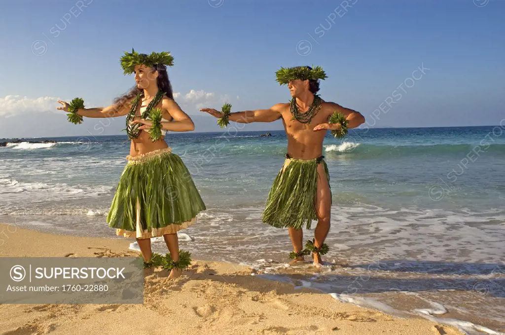 Male and female hula dancers in ti-leaf, haku, lei, in a dancing pose on the beach