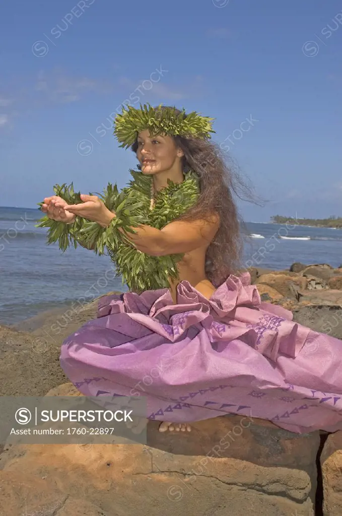 Hula dancer with haku lei in traditional outfit on rocky coast, ocean background