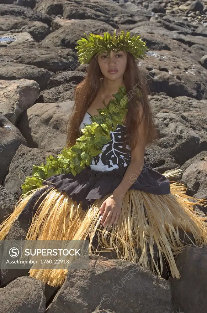 Hula dancer with haku lei in traditional outfit on rocky coast.