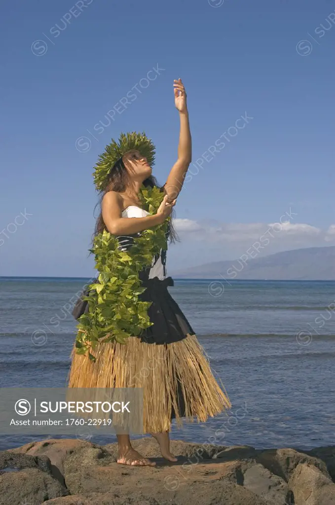 Hula dancer with haku lei in traditional outfit on rocky coast, ocean background