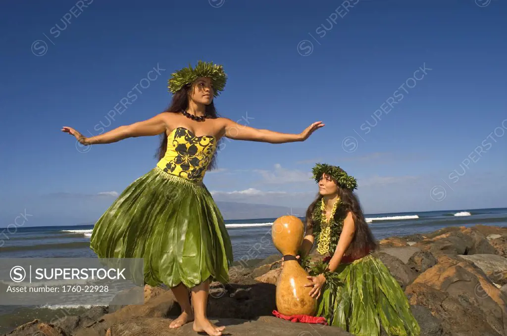 Two hula dancers in traditional outfits on rocky shore, one dancing while the other plays ipu.