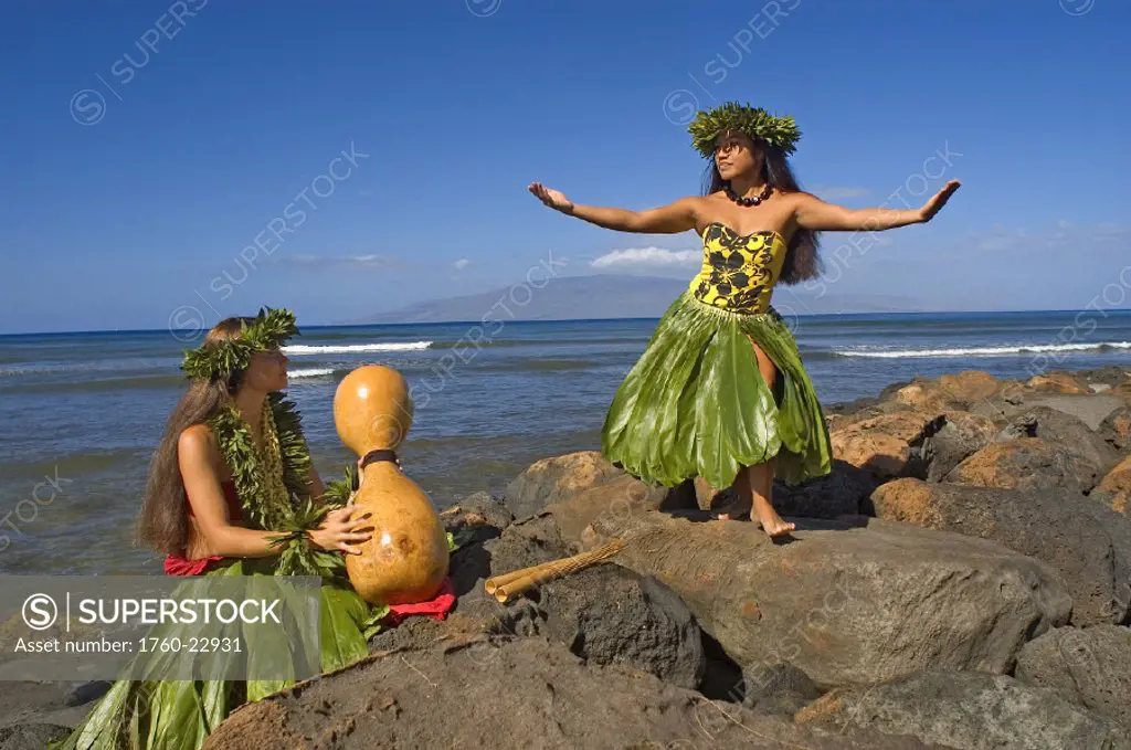 Two hula dancers in traditional outfits on rocky shore, one dancing while the other plays ipu.