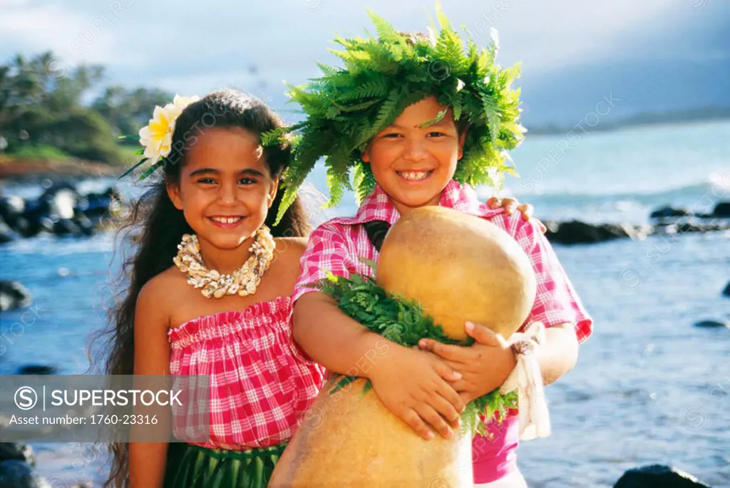 Closeup of two smiling keiki on beach, both wear palaka, boy holds ipu.
