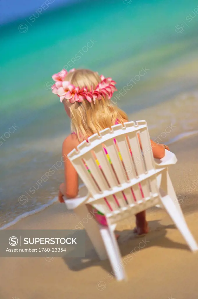 Young blonde girl with haku lei sits in white chair by turquoise ocean, back view