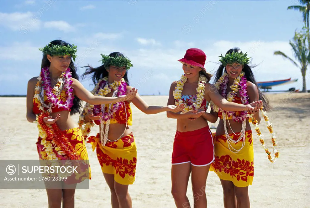 Three Hawaiian women teach visitor to dance hula, all wear leis & smiling, beach