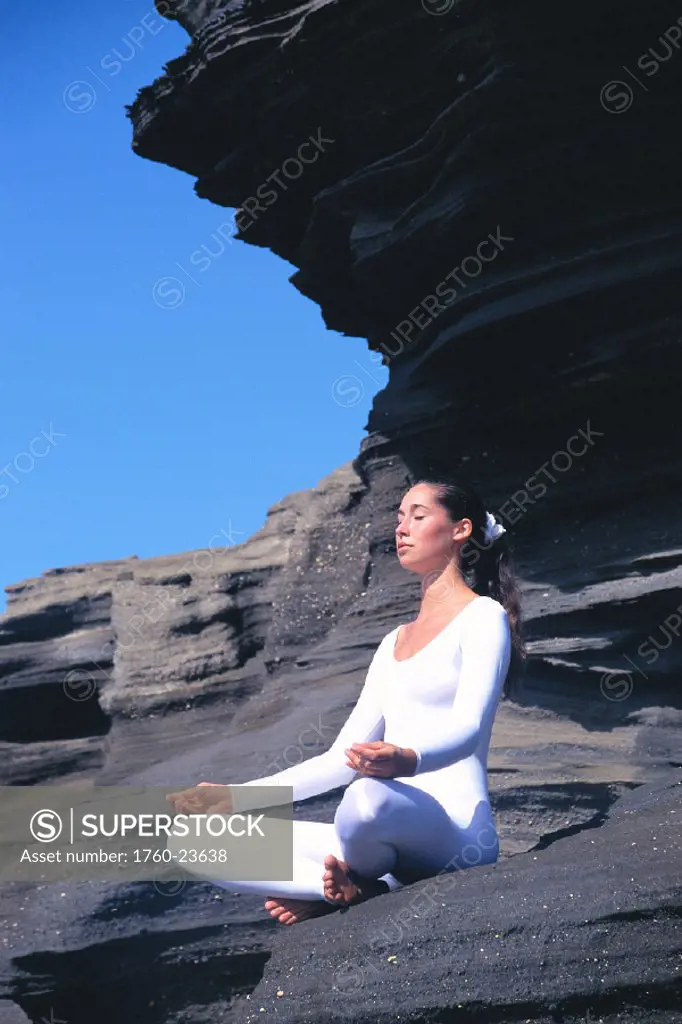 Young woman in white leotard sits, meditating on rocky landscape, yoga C1200