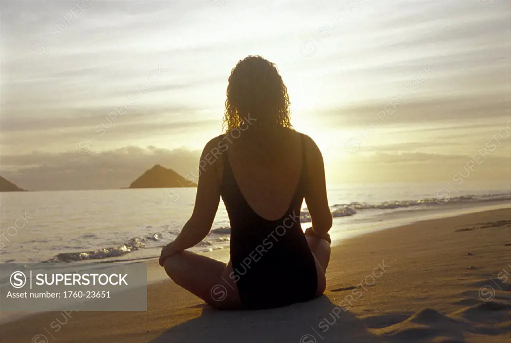 Vu from behind woman sits on beach meditating, facing sunrise D1156 bright golden sky