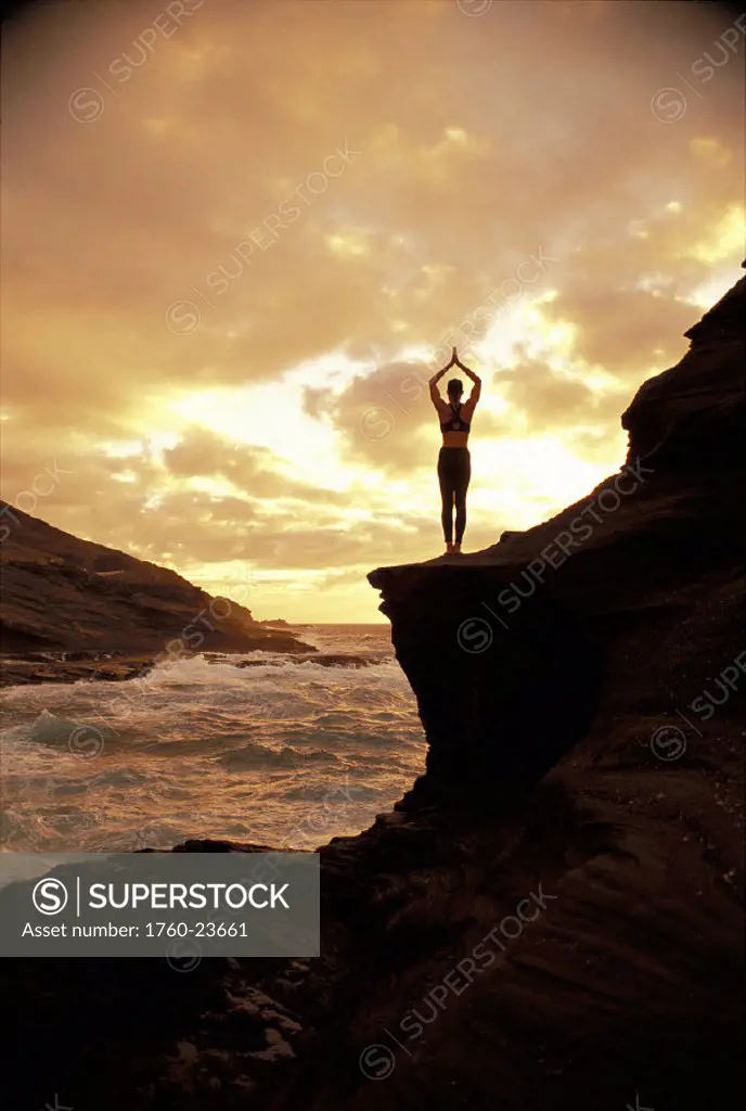 Woman on cliff yoga view from behind turbulent ocean golden pinkish sky D1178