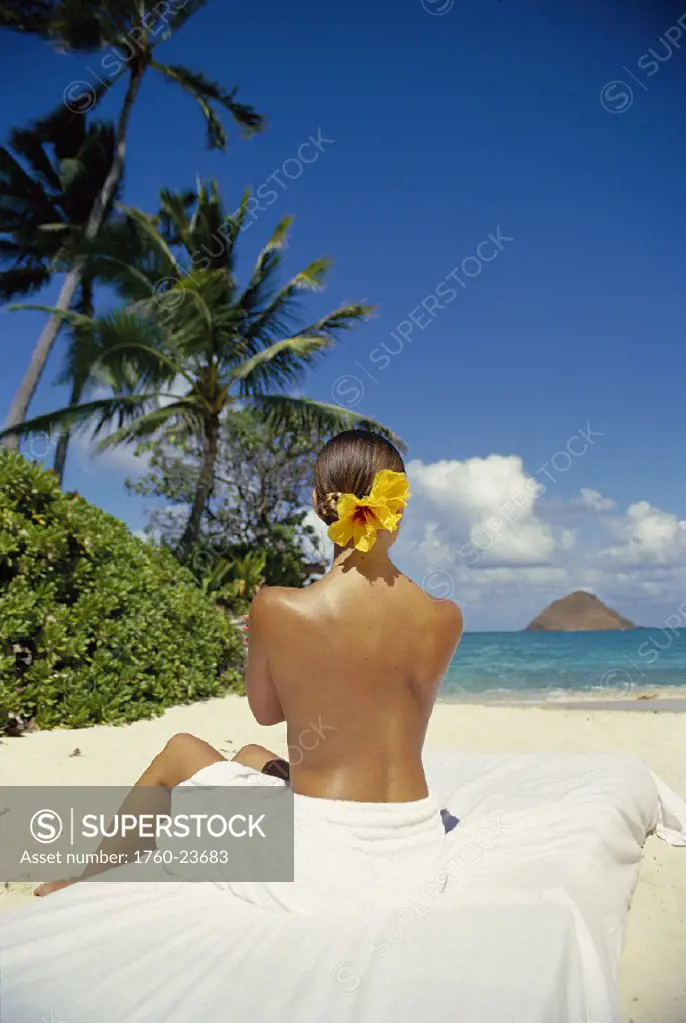 Back vu of woman sitting on white towel @ beach, looking toward ocean, tropical setting