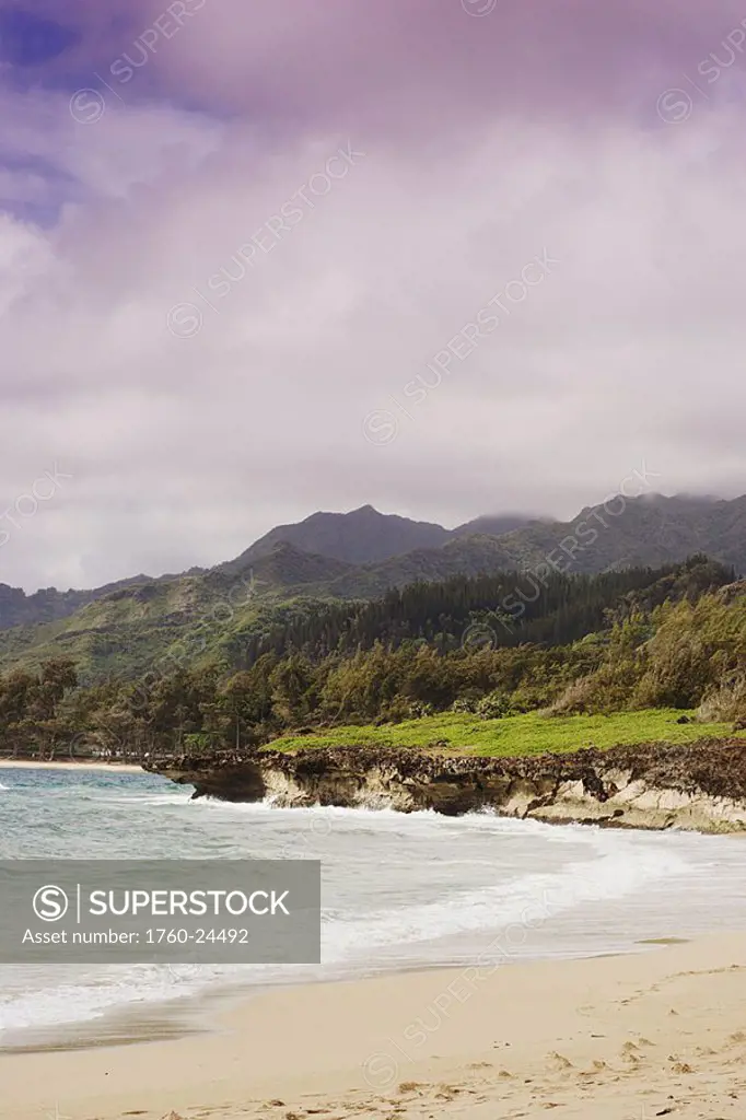 Hawaii, Oahu, Windward, water washes onto sandy beach, lush mountains in background, cloudy sky