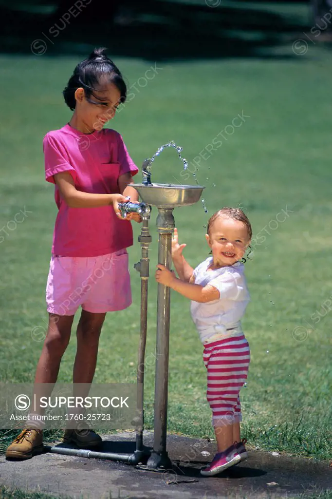 Two girls at water fountain playing, little blonde girl gets wet, smiling C1136