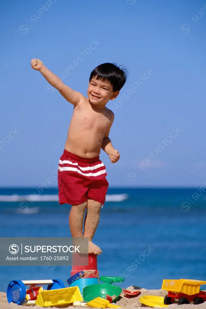 Little boy stands on bucket, smiling with arm up, surrounded with beach toys