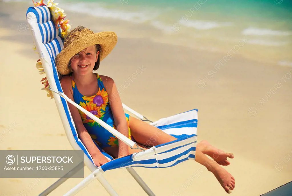 Smiling girl wearing hat sits on beach chair along shoreline, lei draped over