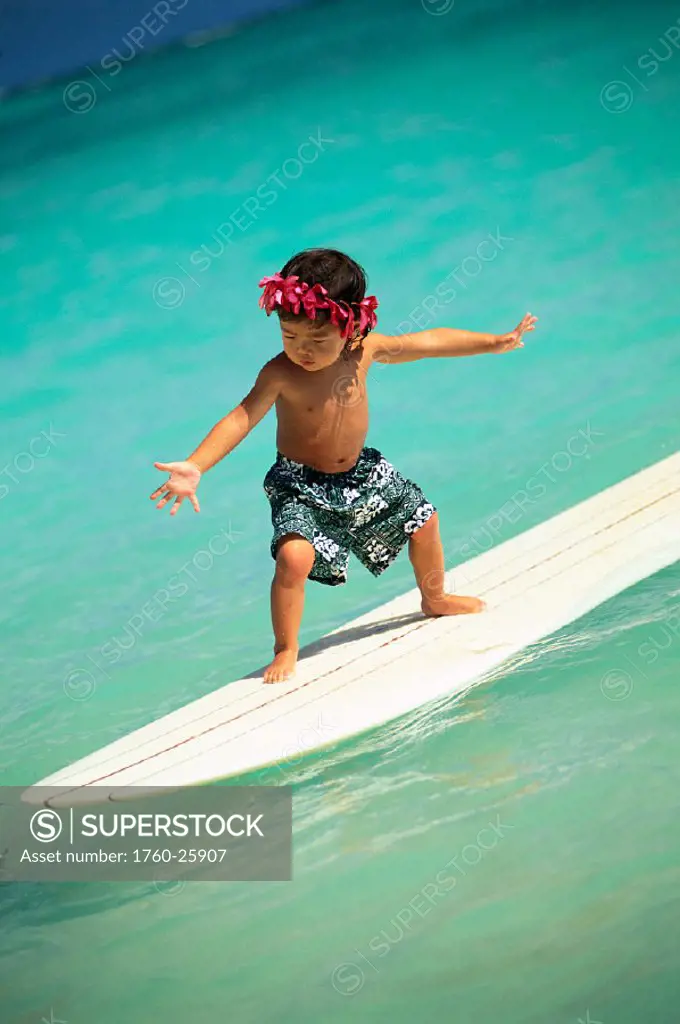 Closeup of little local boy on white surfboard, arms spread, pink haku, turquoise water