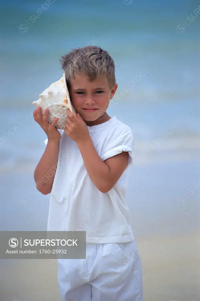 Young boy in white outfit, listening to shell, ocean in background C1056