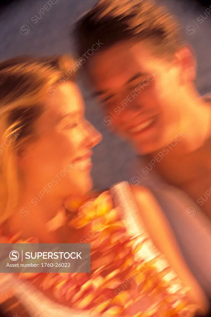Blurred image of a smiling couple, faces only, woman w/ lei, golden lighting