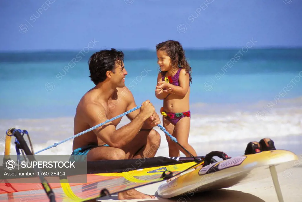Father and young daughter on beach with windsurf gear.