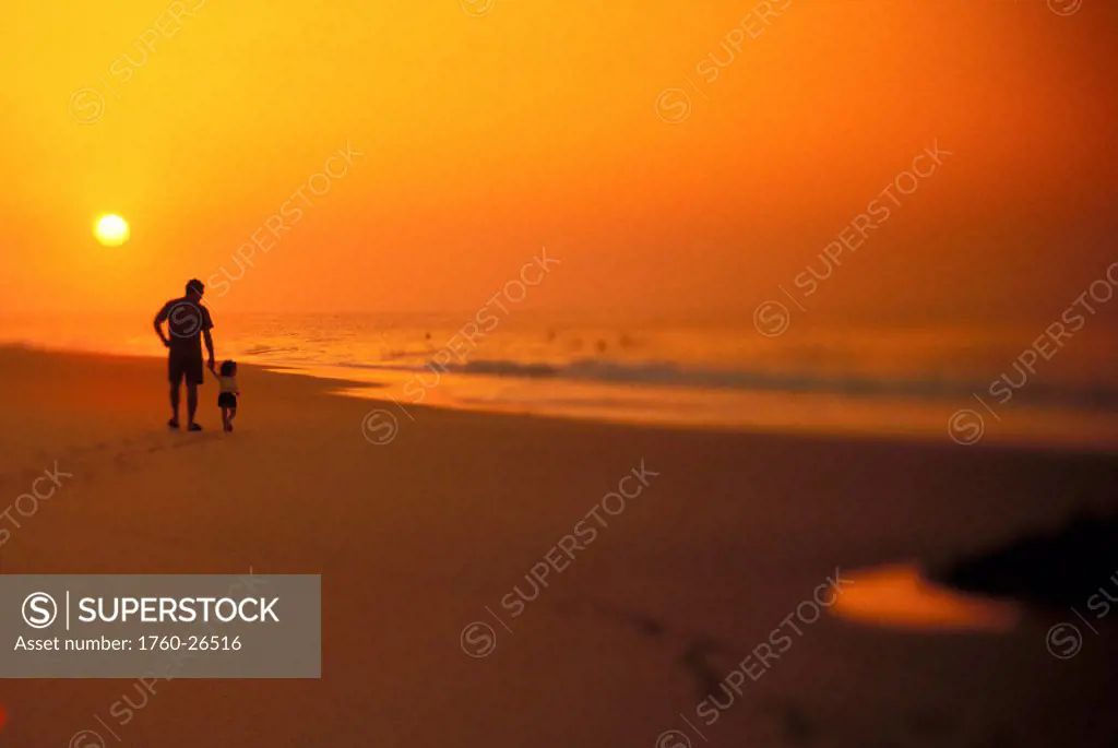 Distant view of father and son walking along shoreline, fiery sunset