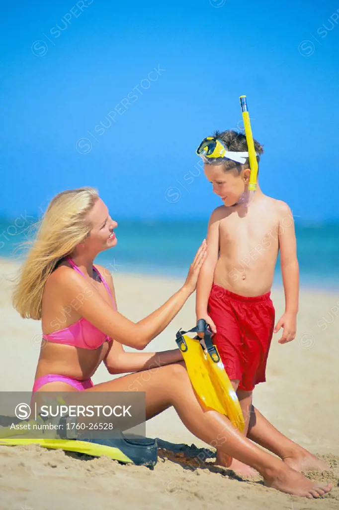 Mother and son on beach in colorful swimwear, relaxing w/ snorkel gear, blue C1081 sky