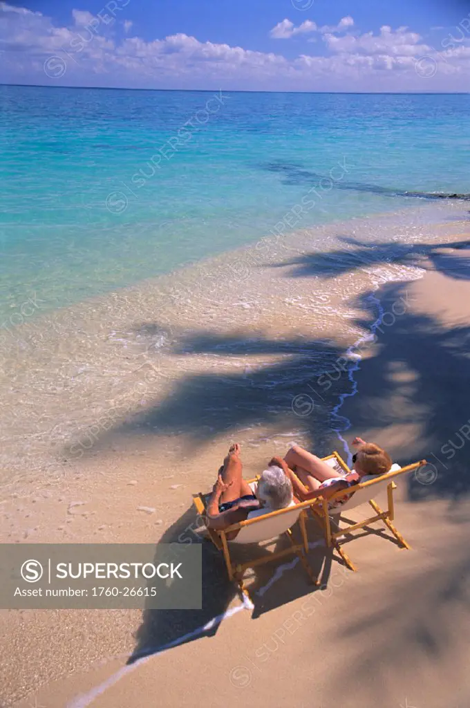 Vu from above, senior couple in lounge chairs along shore break, turquoise water, palm shadows