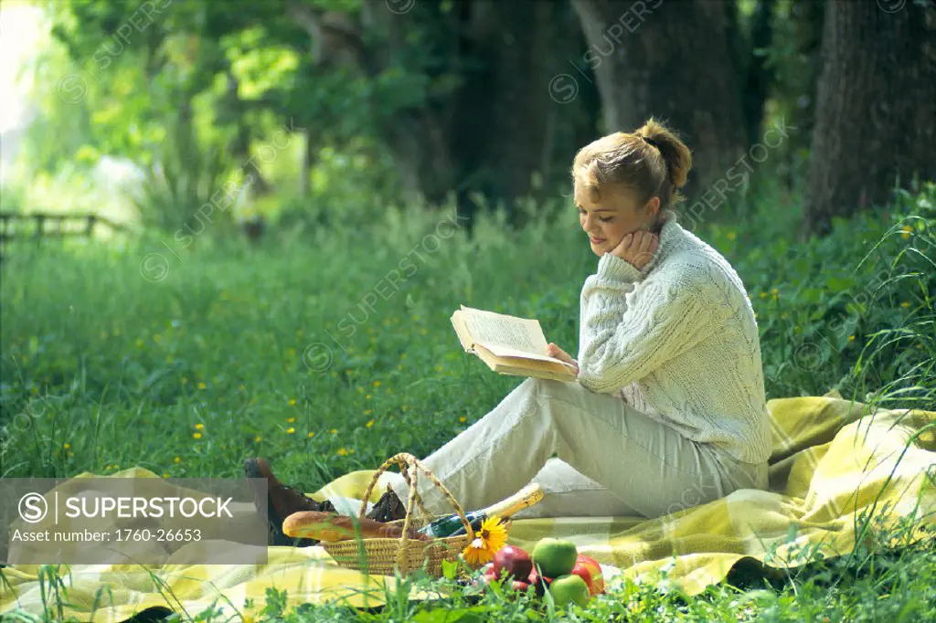 Woman reading book in country setting with picnic basket, fruits C1143
