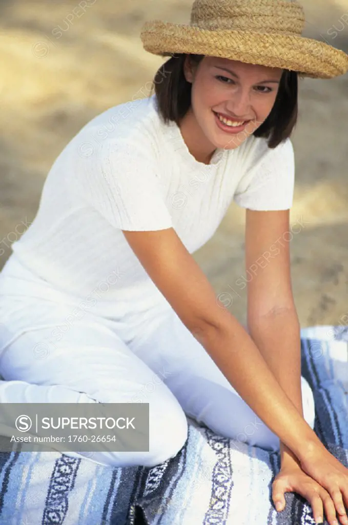 Smiling woman with hat sitting on blanket at beach, leaning forward