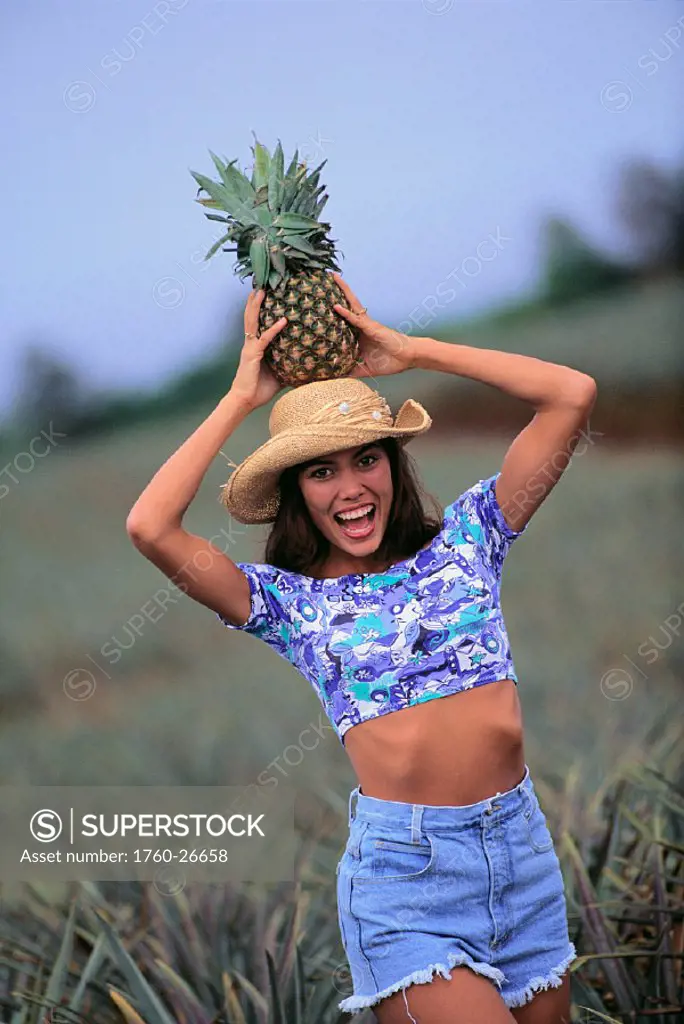 Excited young woman wearing hat, holding pineapple atop head, in fields