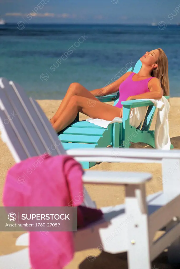 Woman lounging with eyes closed, beach chair w/ towel in foreground, ocean bkgd