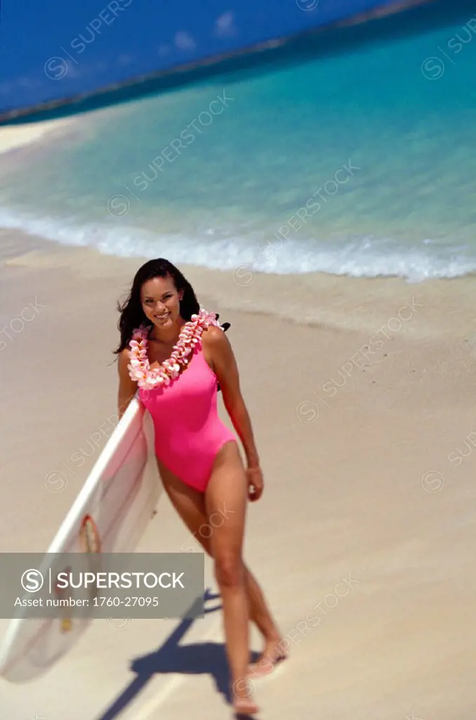 Woman on beach with surfboard, walking, smiling, blue sky, wears lei