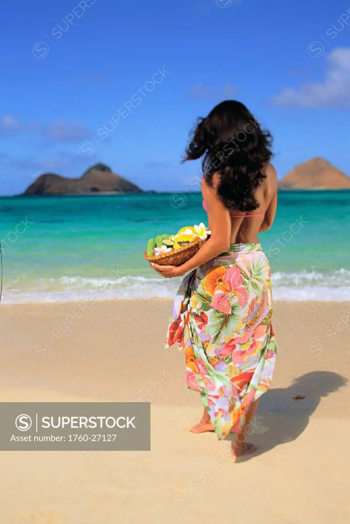 Back view of beautiful woman walking toward turquoise water holding fruit basket