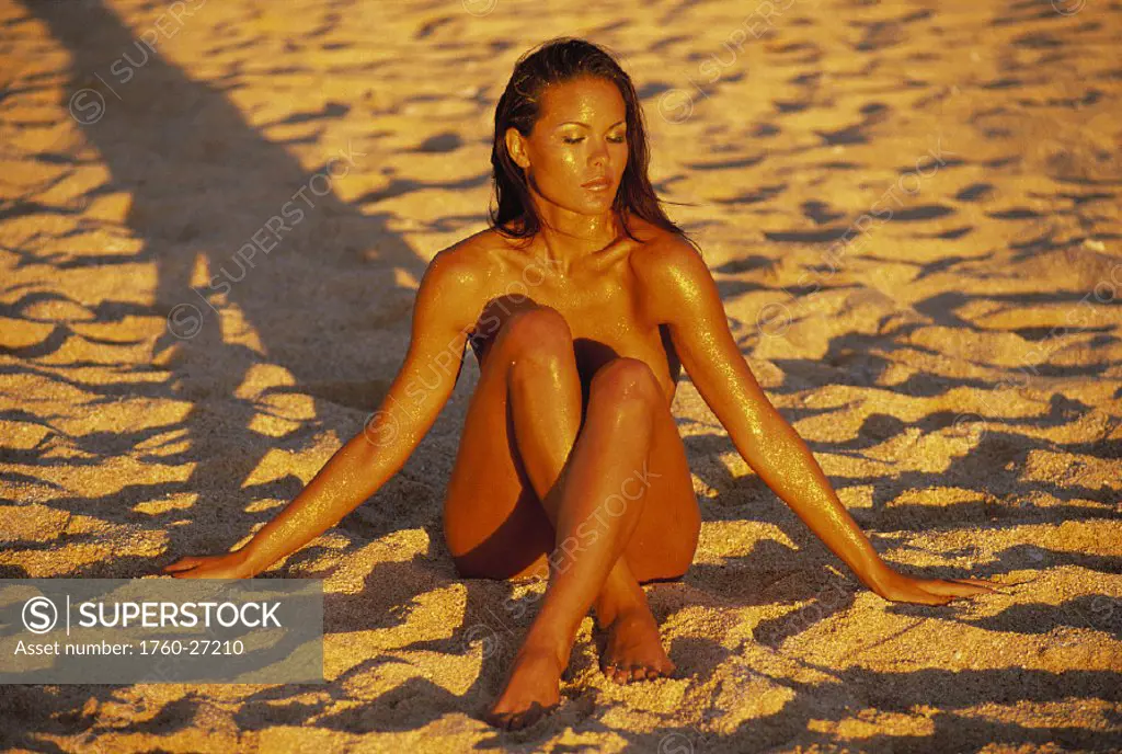 Sexy woman sits on beach bare w/ legs crossed hands in sand golden shimmery, shadow naked