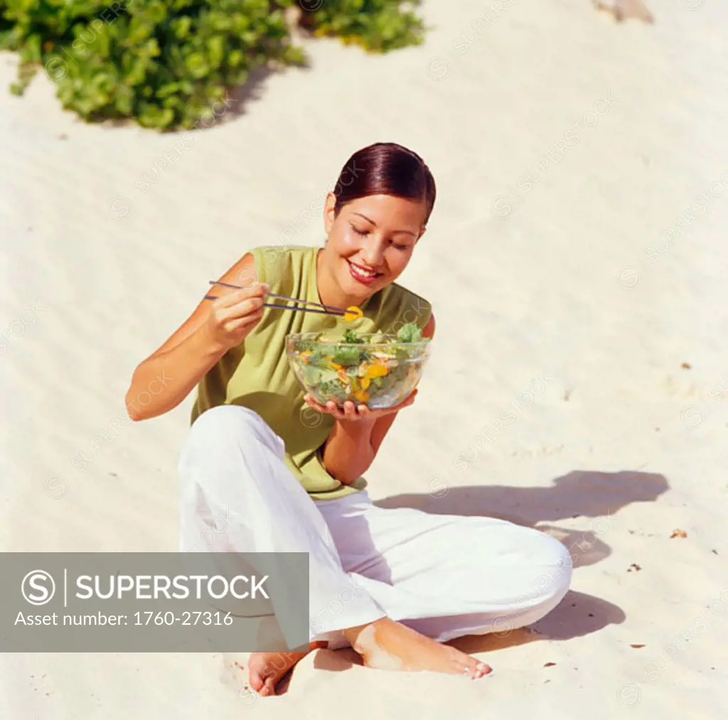 Young woman eating a salad with chopsticks on the beach.