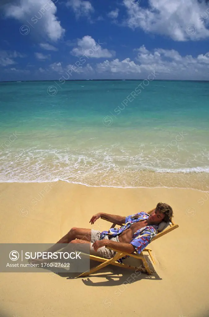 Man in aloha shirt relaxes in beach chair along shoreline waters