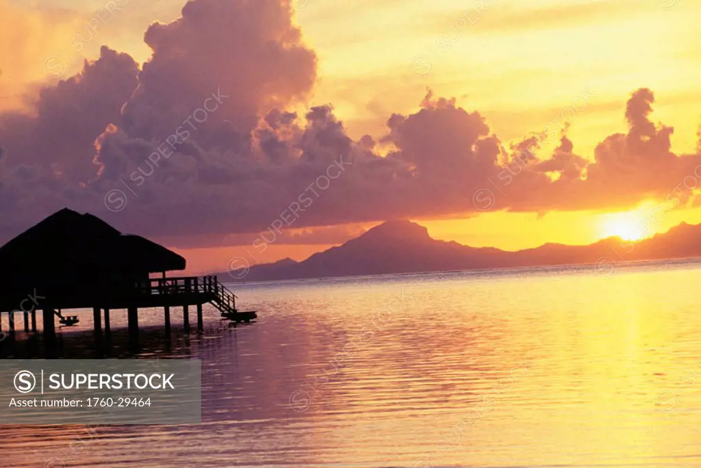 French Polynesia, Tahiti, Huahine, Te Tiare Resort bungalow over ocean, golden sunset with reflections, Raiatea in background