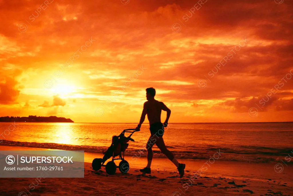 Guam, Tumon Bay, Man with baby stroller silhouetted at sunset, jogging on beach