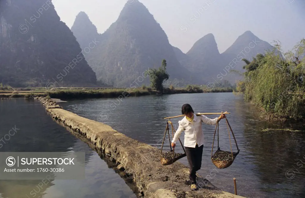 China, Yangshuo, woman carries baskets along manmade walkway across river, mountains in background