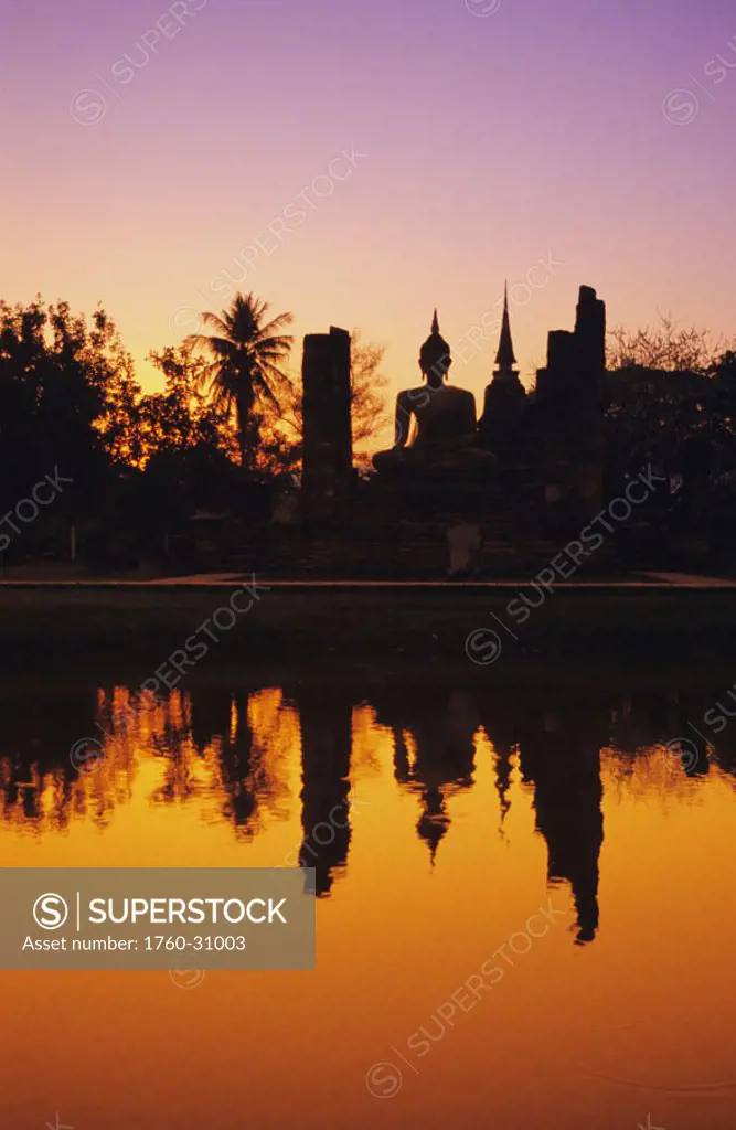 Thailand, Sukhothai, Wat Mahathat, distant view of Buddha statue at sunset, silhouetted and reflecting on water.