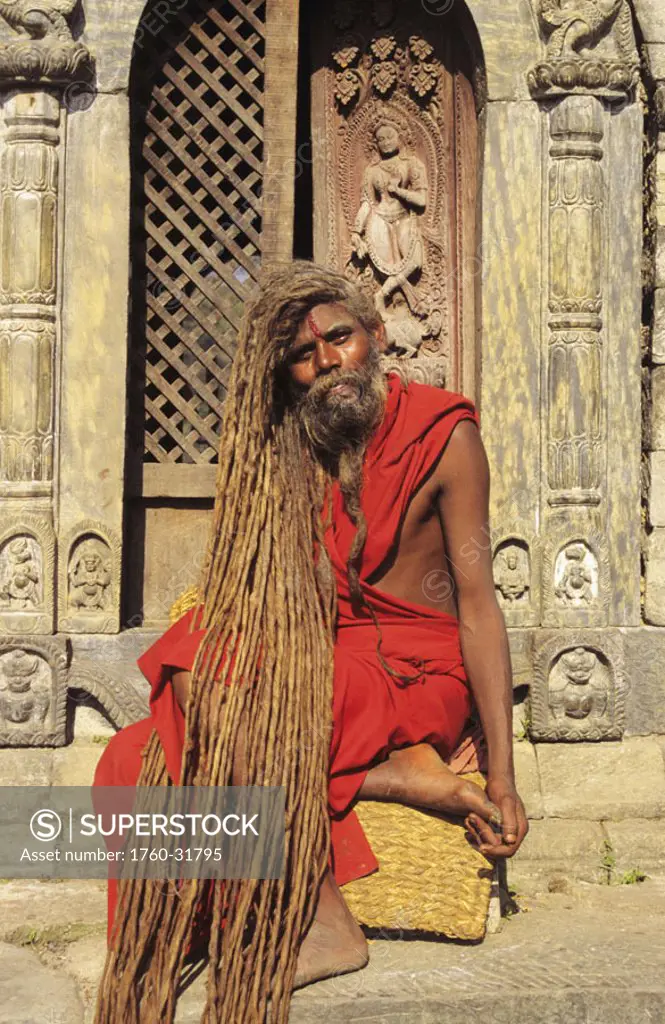 Nepal, Kathmandu, Pashupatinath Temple, Hindu holy man sitting on stairs wearing red robe, long dreadlocks.