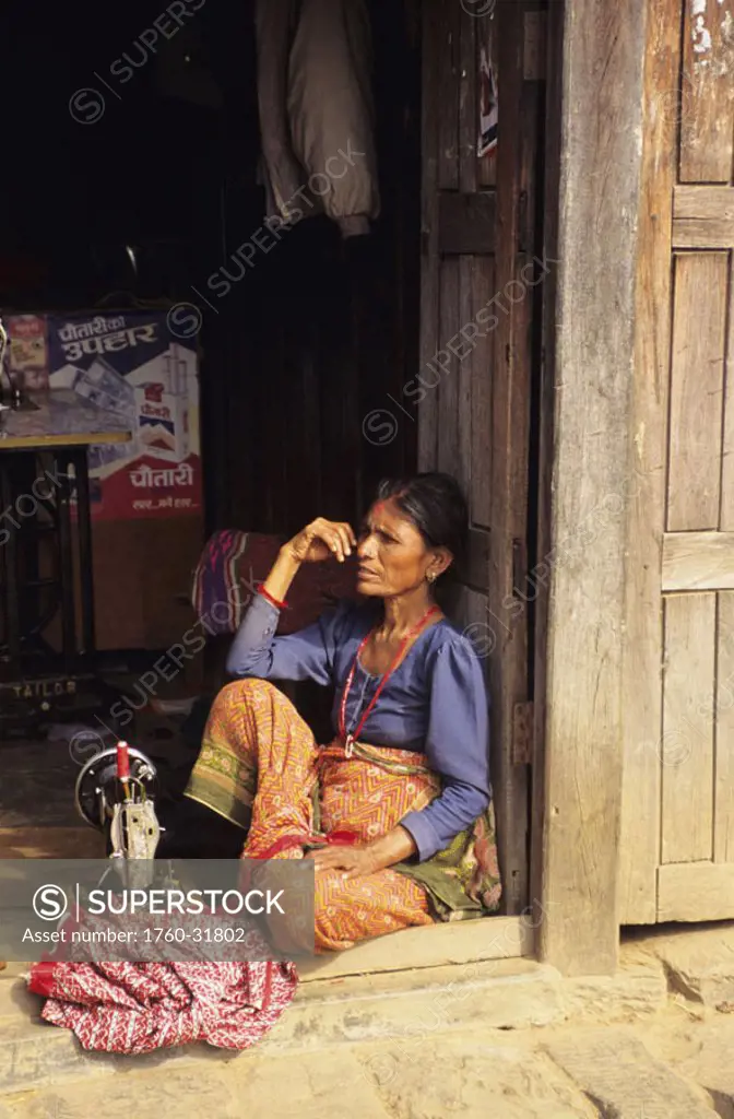Nepal, Gorkha, village woman sitting on doorstep, sewing machine on floor with clothing.