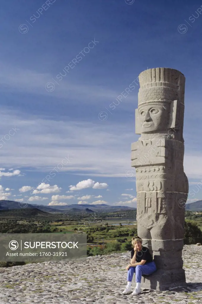 Mexico, Tula, Toltec Ruins, senior woman sitting at base of stone sculpture, country landscape in background.