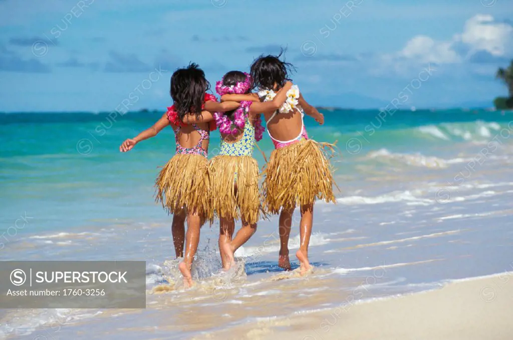 Back view of three young girls on beach, grass skirts, lei, arms around each other, run in water