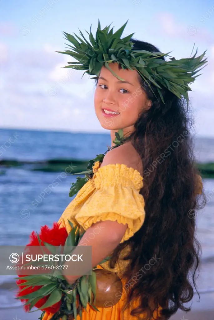 Portrait shot, side view of young girl in hula attire on beach smiling, hands C1457 on hips