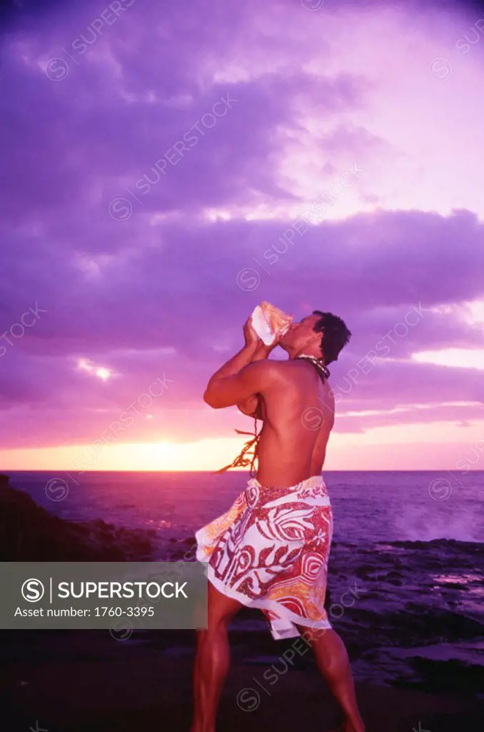 Hawaiian man blows conch shell at beach, sunrise with purple and yellow sky.